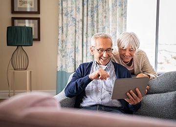 A photo of a senior couple finding peace of mind in genuine customer reviews following hernia surgery. A gent is sat on a sofa holding an iPad as his companion looks over his shoulder.