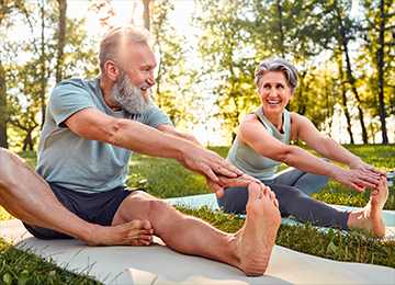 This photo shows to seniors gently stretching their legs, while sat outside on green grass in the morning sun. They are smiling.