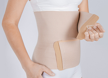 This photo shows a torso with a beige support that is snuggly wrapped around the waist and rib cage. The wearer holds a second fixing, ready to Velcro the support in place.