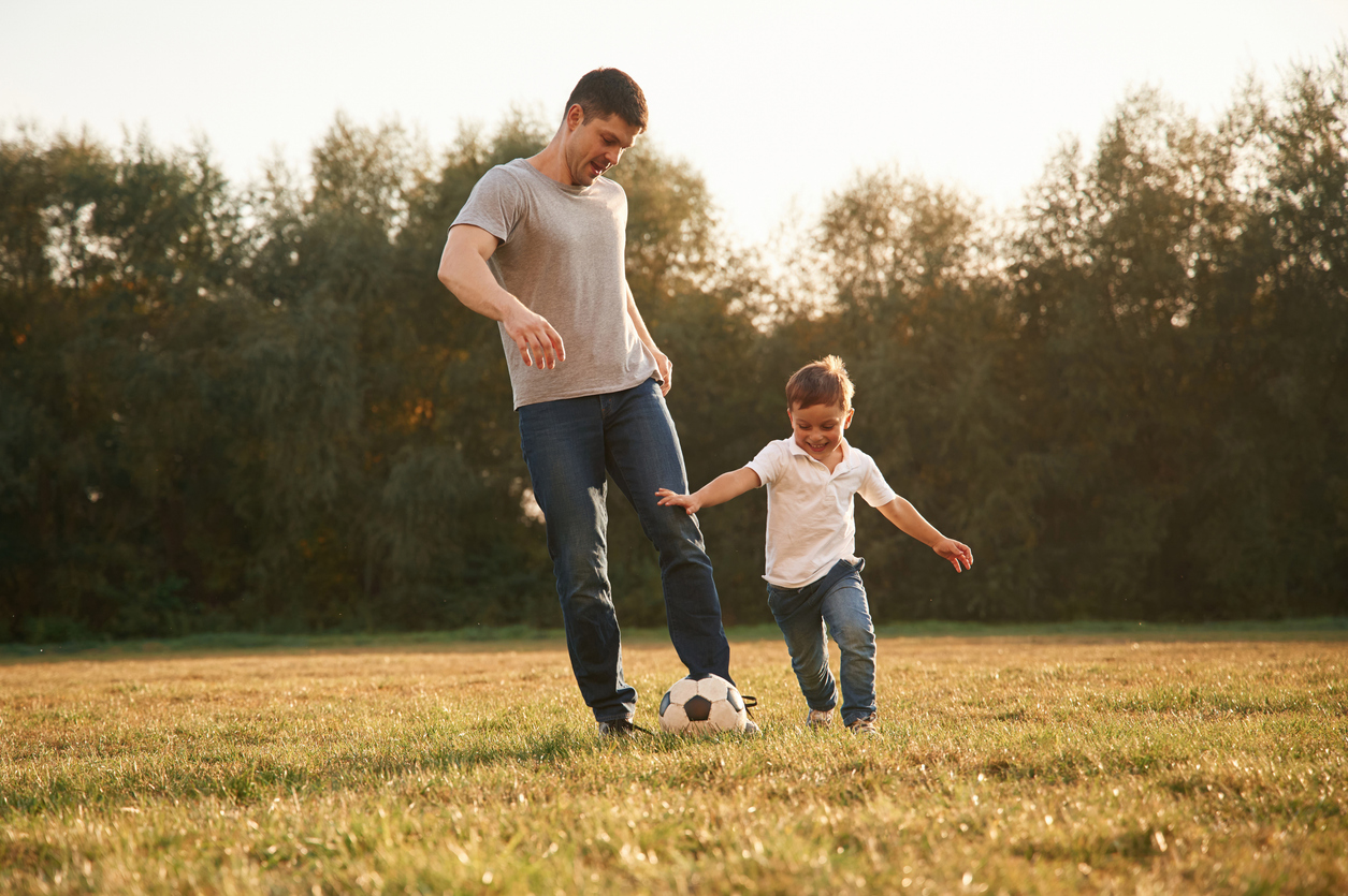 A man kicking a football alongside a young child in a field, not held back by a sports hernia/Gilmore's groin.