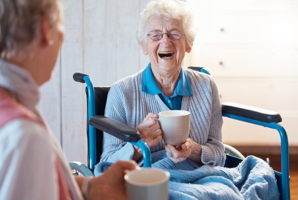 Senior women recovering from hernia surgery, sitting in a wheelchair and holding a mug of tea, laughing with a friend.